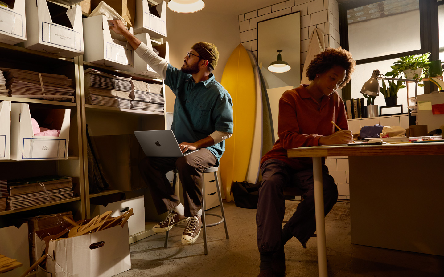 Osoba siedząca przy biurku pisze ołówkiem w notesie, a inna – z MacBookiem na kolanach – sięga prawą ręką po teczkę z półki.