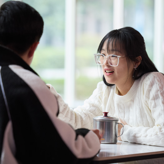 Two people sit facing each other at a cafeteria table and talk