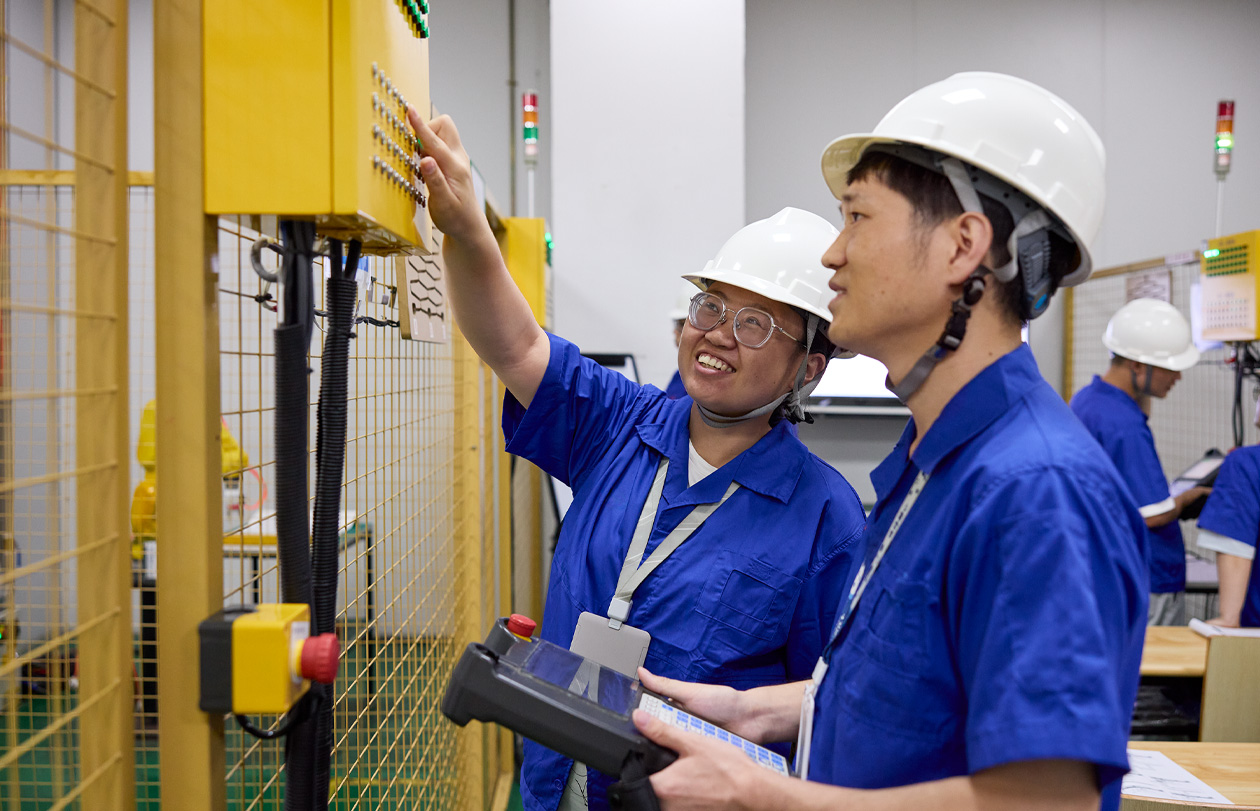 Two employees wearing safety hats and uniform shirts stand by a work cage control box, where one points out a button to the other